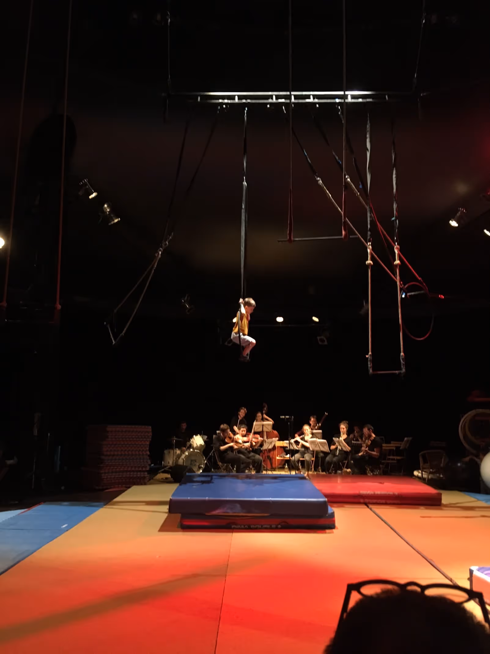 Enfant en suspension aérienne sur des cerceaux au-dessus de l'orchestre et du tapis multicolore, spectacle de l'école du Cirque Électrique