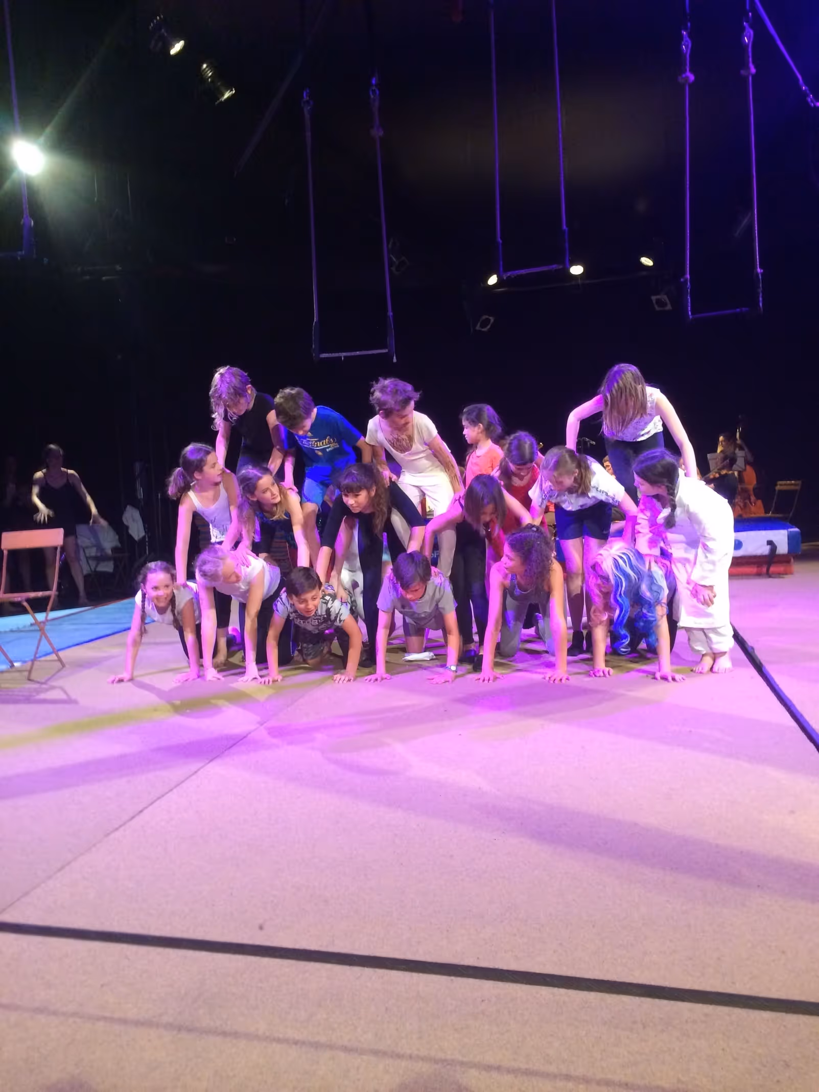Groupe d'enfants formant une pyramide humaine sous éclairage violet avec trapèzes suspendus au-dessus, spectacle de l'école du Cirque Électrique