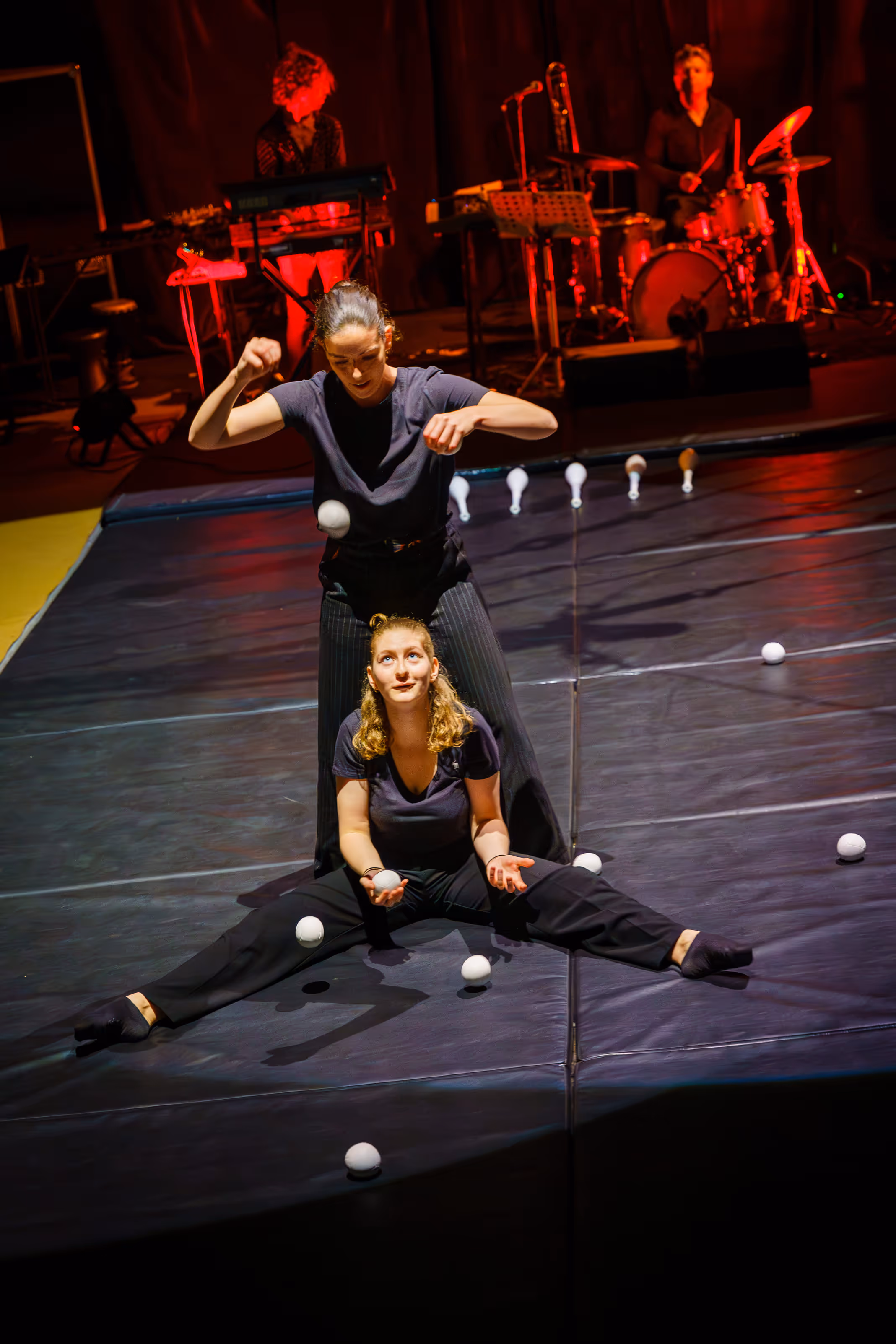 Deux femmes en noir jonglant avec des balles blanches sur une scène, avec un groupe de musiciens en arrière-plan éclairé en rouge.