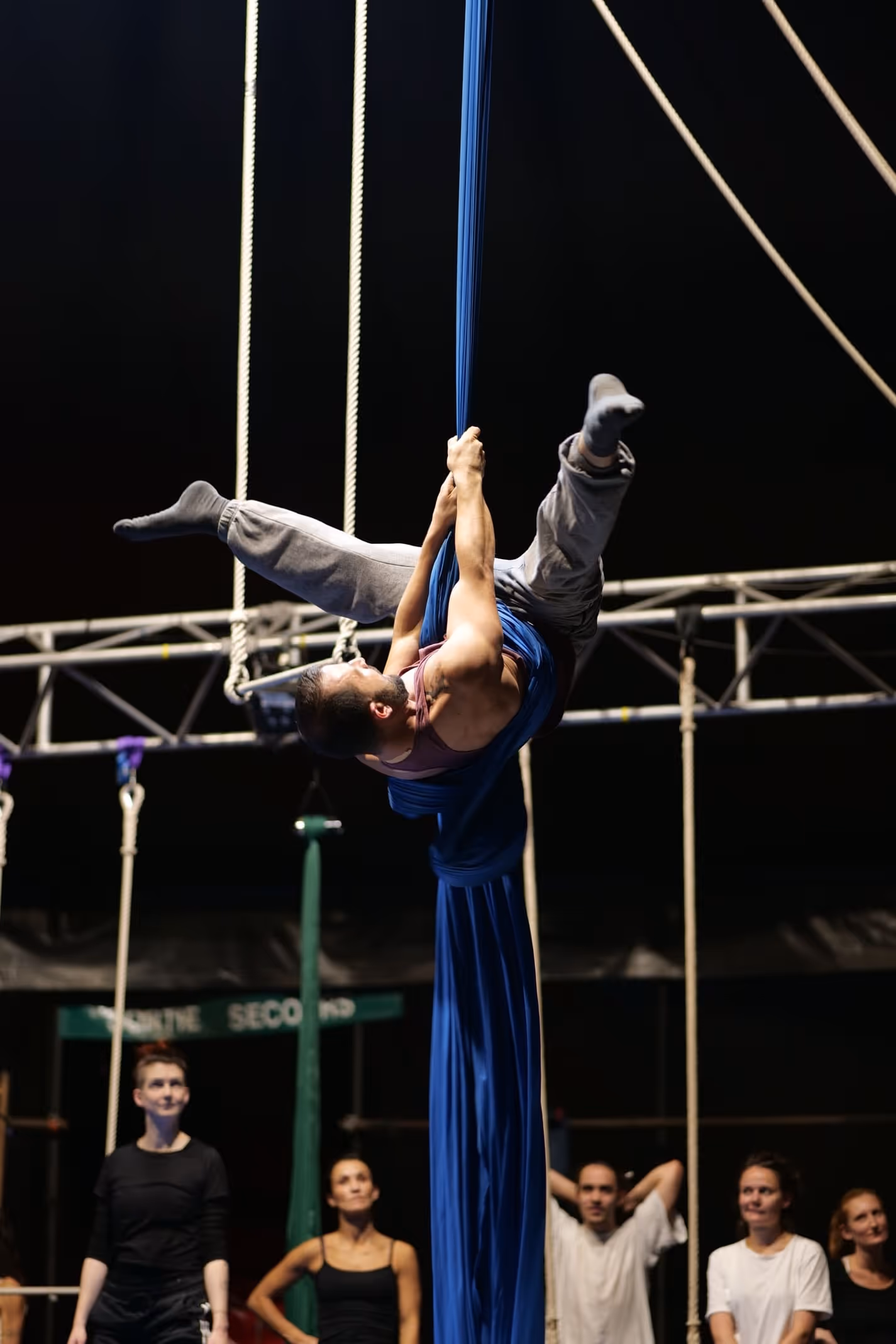 Un homme effectuant une acrobatie suspendue avec un tissu bleu devant un groupe de spectateurs attentifs.