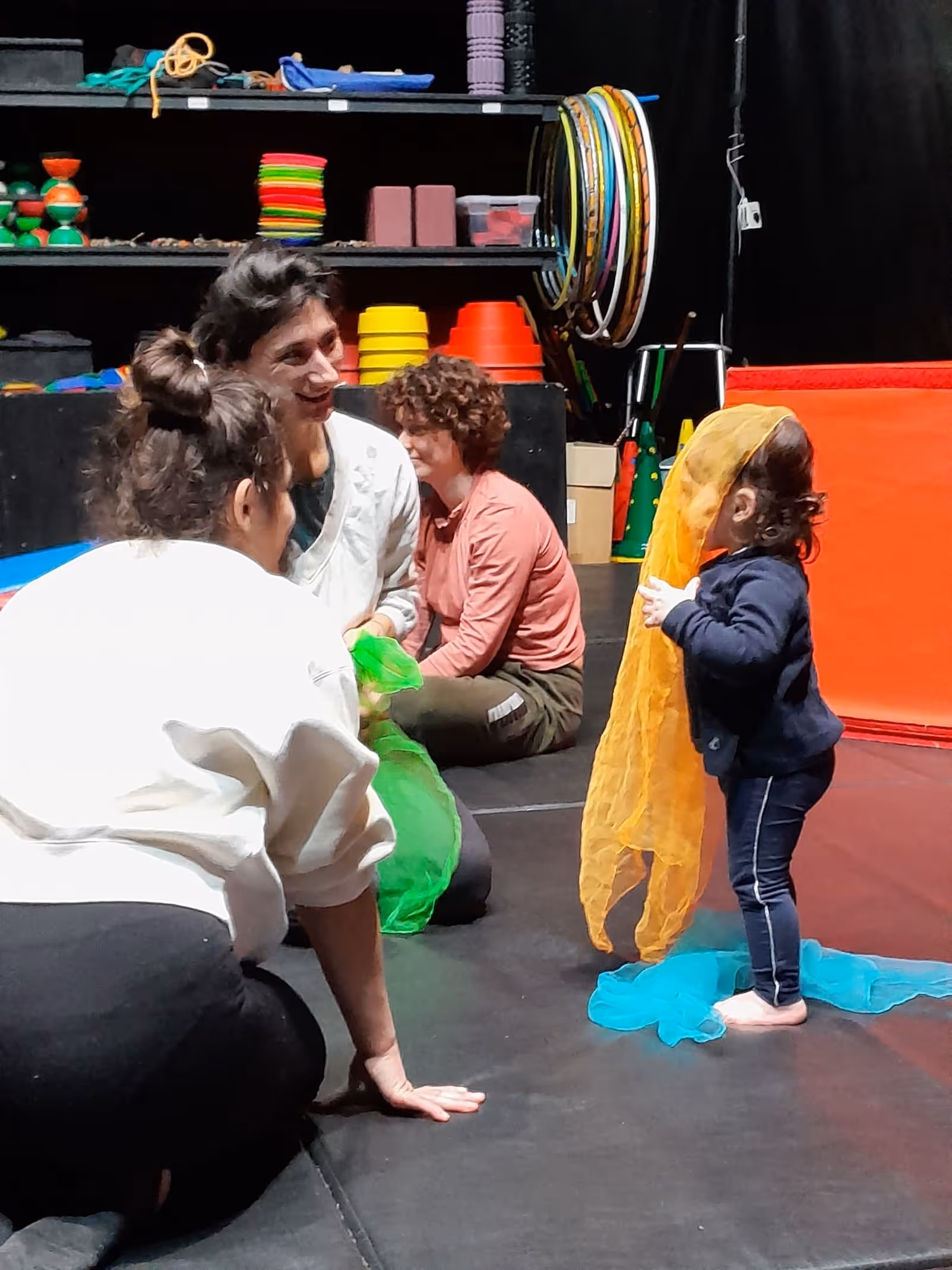 Une petite fille joue avec un foulard orange tandis que trois adultes l'observent et sourient dans une salle avec des équipements colorés en arrière-plan.