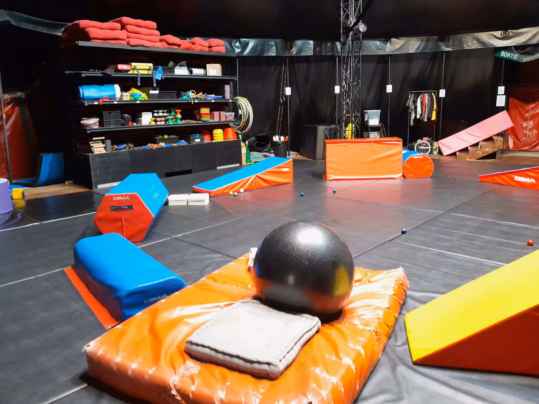 Salle d'entraînement de cirque avec tapis amortisseurs colorés, grande balle noire et étagères avec divers équipements d'entraînement.
