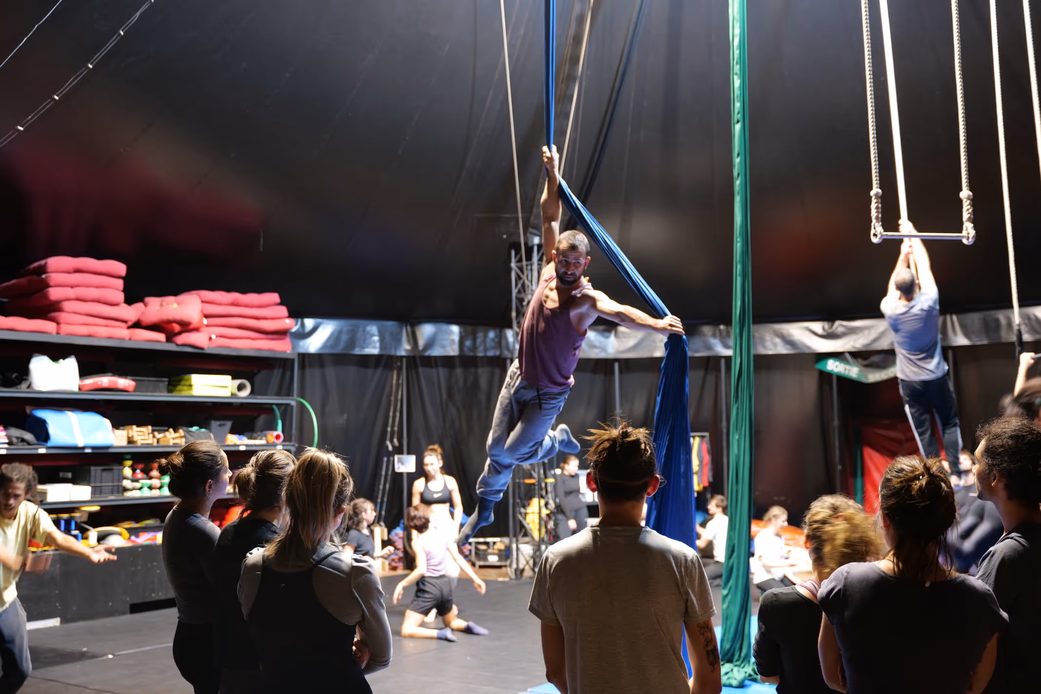 Un homme réalise un exercice aérien sur tissu bleu dans un studio de cirque, avec plusieurs personnes observant et un trapèze suspendu à droite.