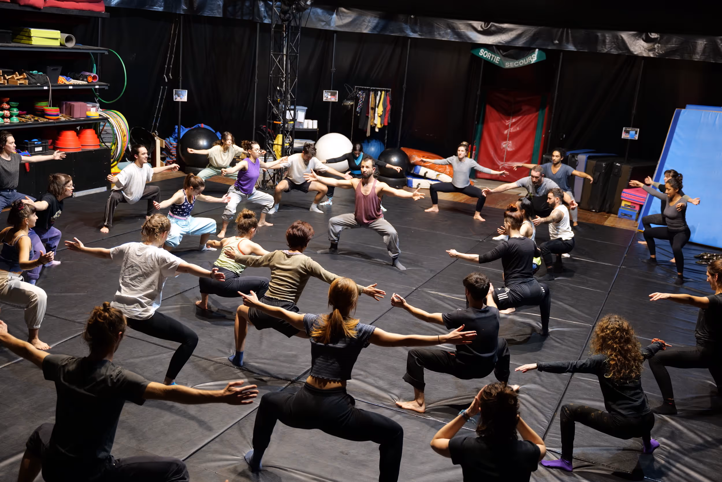 Groupe d'adultes en cercle effectuant des fentes latérales lors d'un échauffement, matériel de cirque visible en fond de salle, cours avec Selwan Cherfi acrobate aérien à l'école du Cirque Électrique.