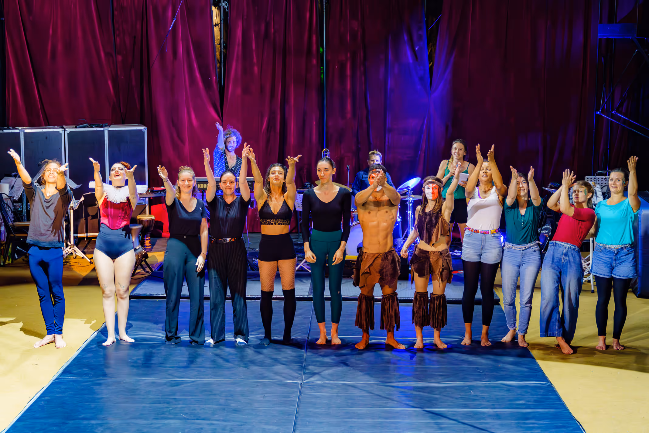 Une troupe de quinze artistes sur scène saluant le public devant un rideau rouge, certains en costumes de spectacle.