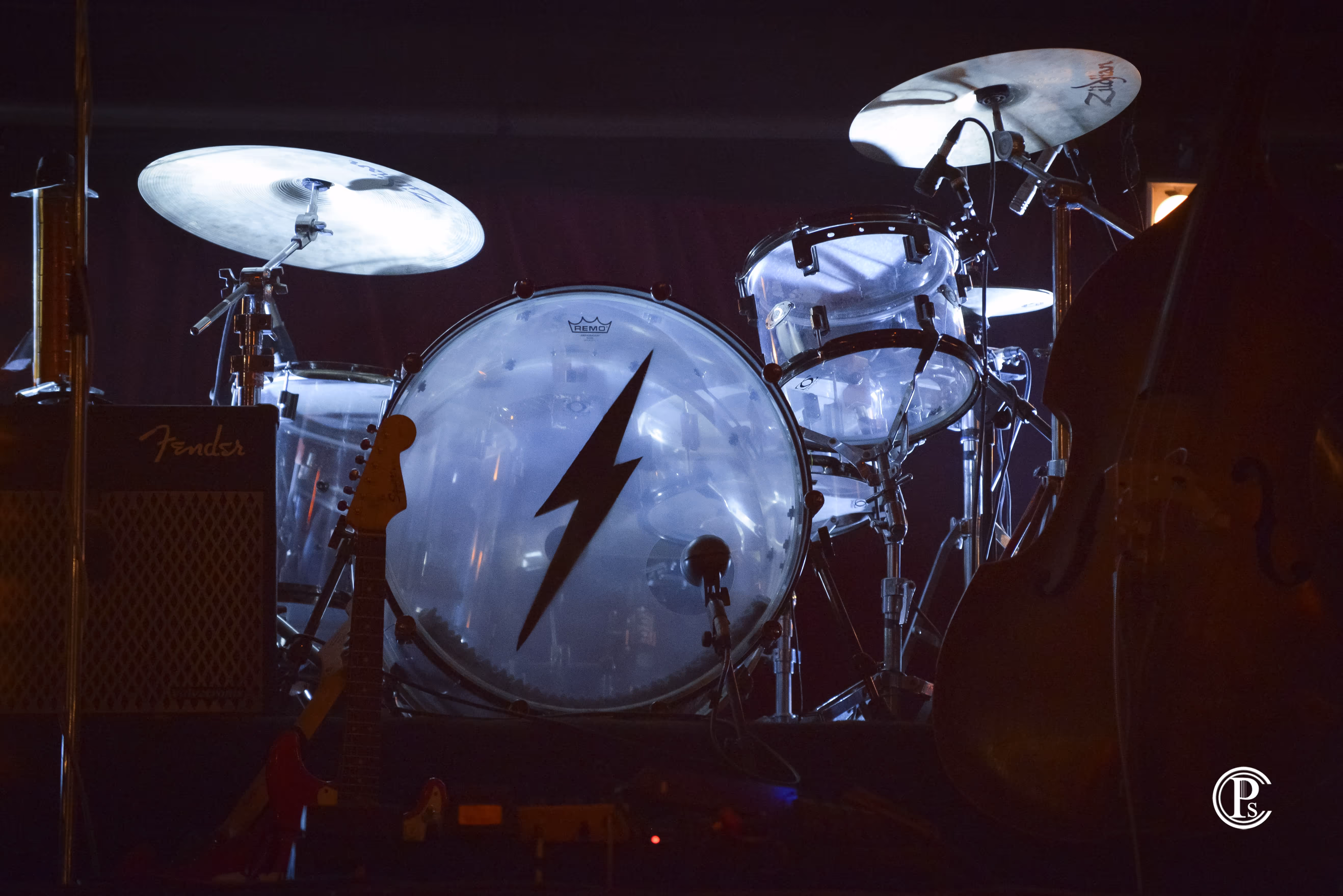 Batterie éclairée sur scène avec symbole d’éclair sur la grosse caisse, guitare électrique et ampli Fender à gauche.