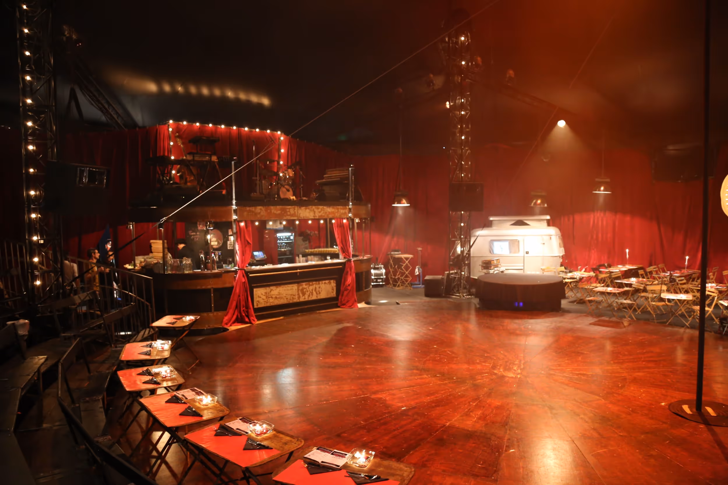 Intérieur d'un chapiteau de cirque avec piste en bois circulaire, tables et chaises pliantes, bar éclairé, et une caravane blanche en arrière-plan.