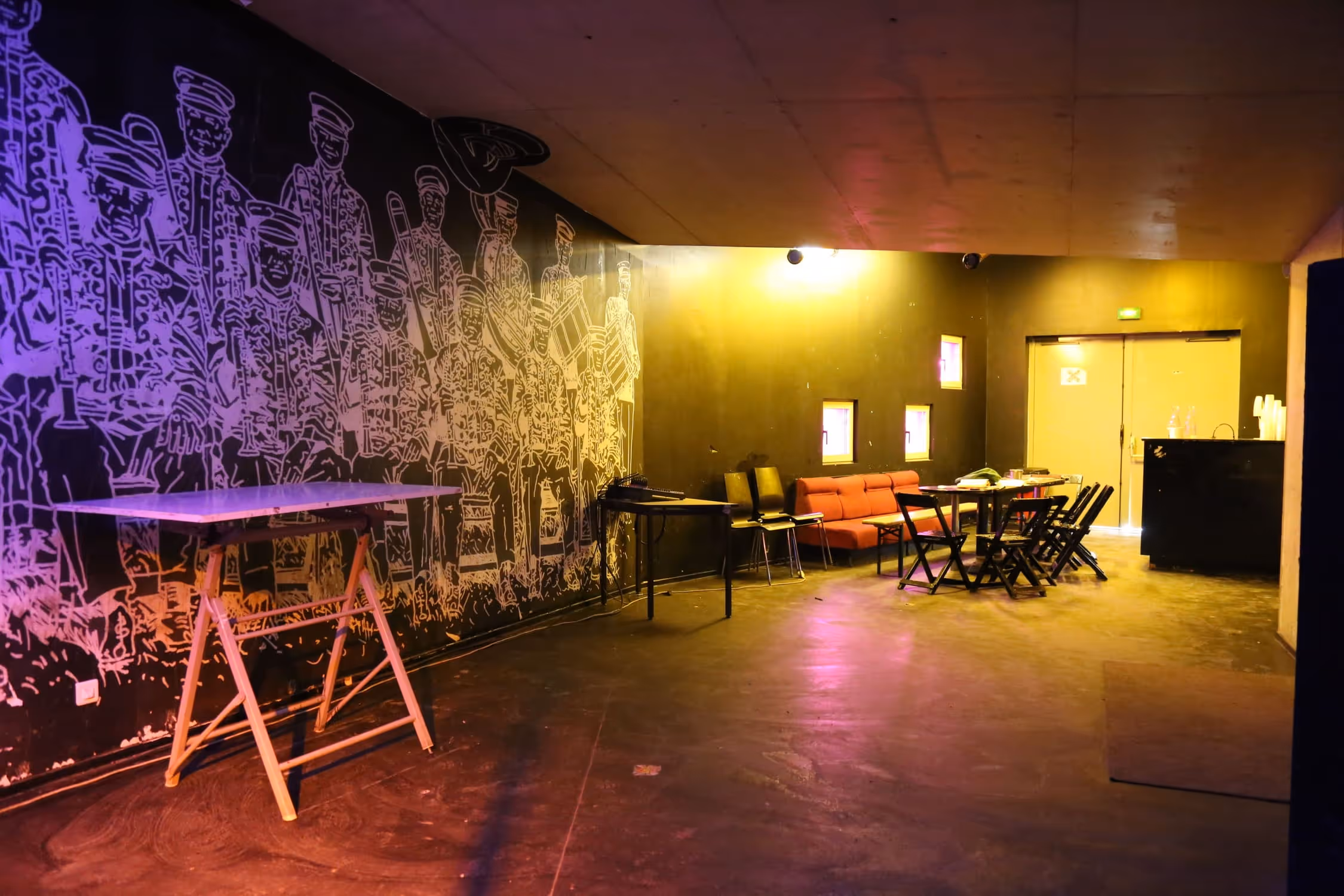 Salle avec un mur peint de silhouettes d'un orchestre, tables pliantes, un canapé rouge, et plusieurs chaises sous un éclairage jaune et violet.