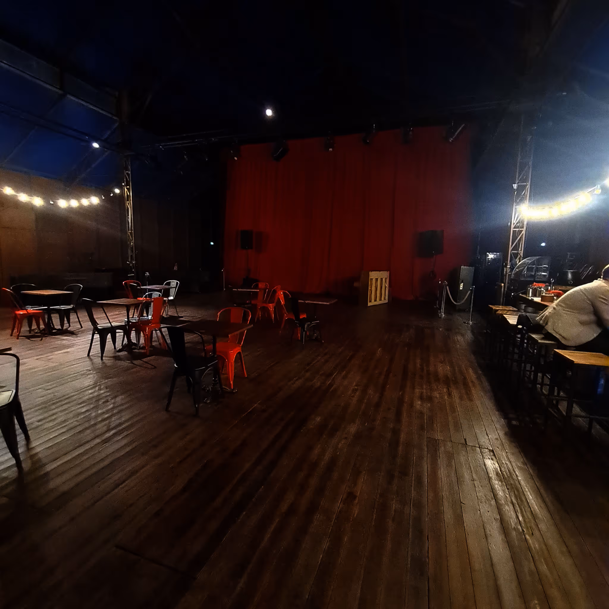 Vue intérieure du parquet de bal avec un sol en bois, des tables et des chaises rouges et noires éparpillées, un mur avec un rideau rouge et un bar avec une personne assise.