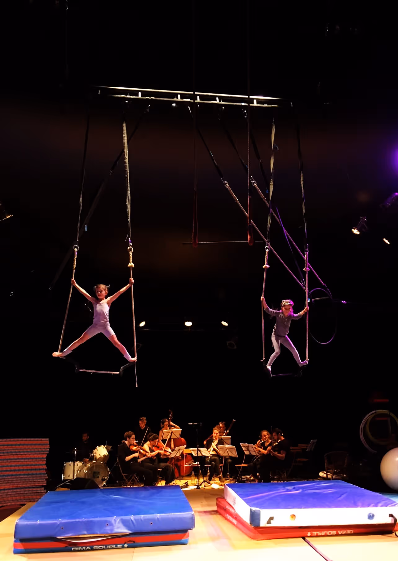 Deux jeunes enfants pratiquent le trapèze suspendu au-dessus de tapis bleus avec un orchestre jouant en arrière-plan sous un éclairage de scène.