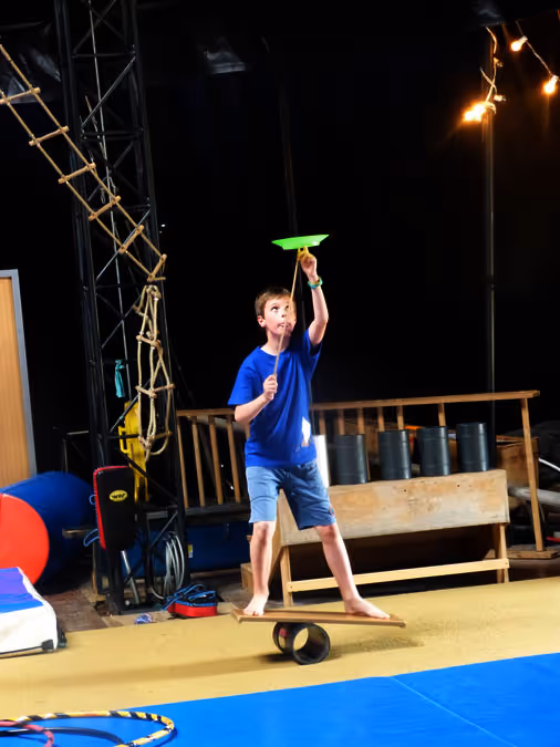 Garçon en t-shirt bleu et short bleu clair, debout pieds nus sur une planche d'équilibre, faisant tourner une assiette verte sur un bâton dans une salle de cirque.