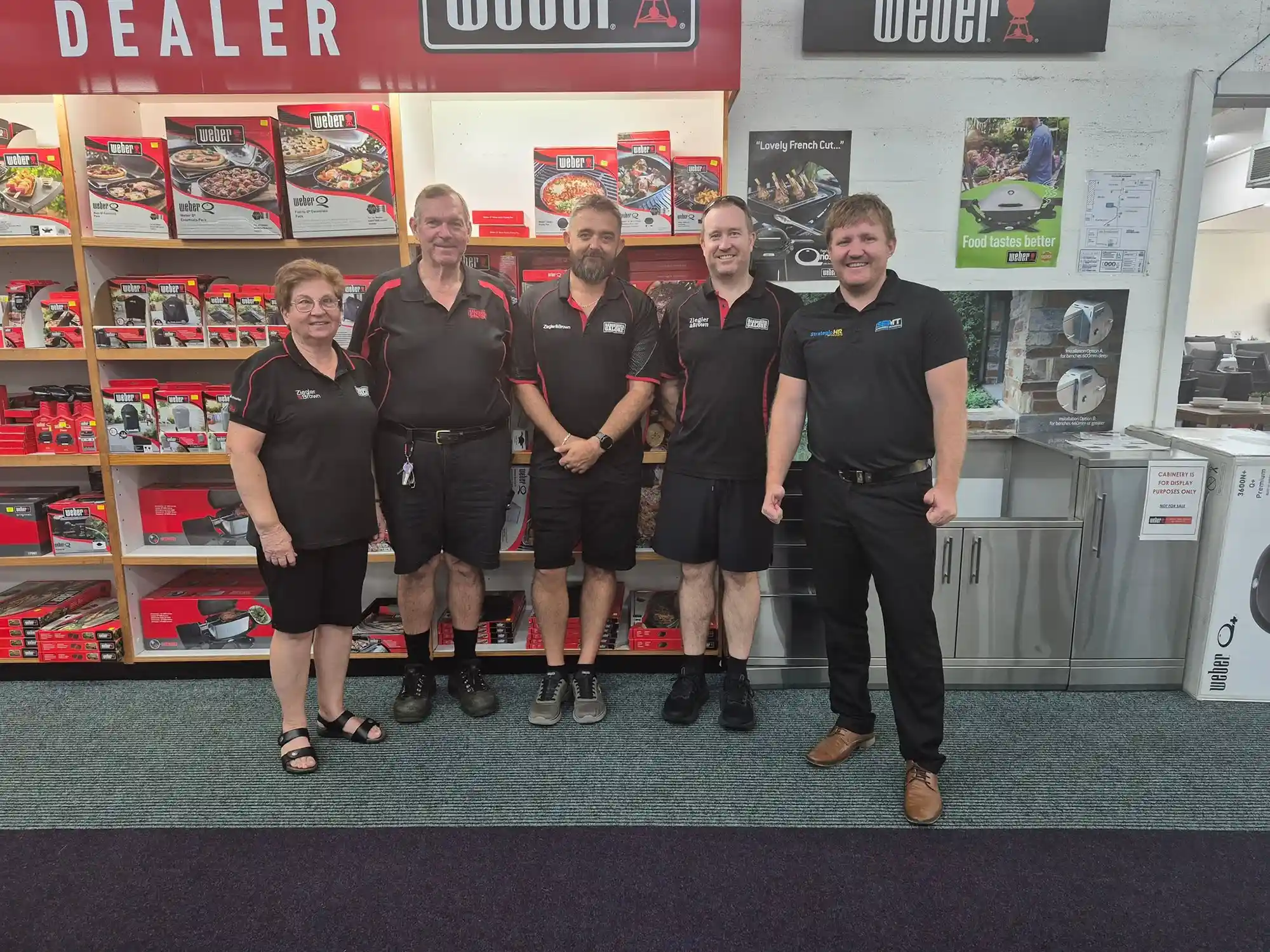 Group photo of the BBQs Galore Team in their store with Nathan from Strategic Business Co in front of a wall of products on the shelf