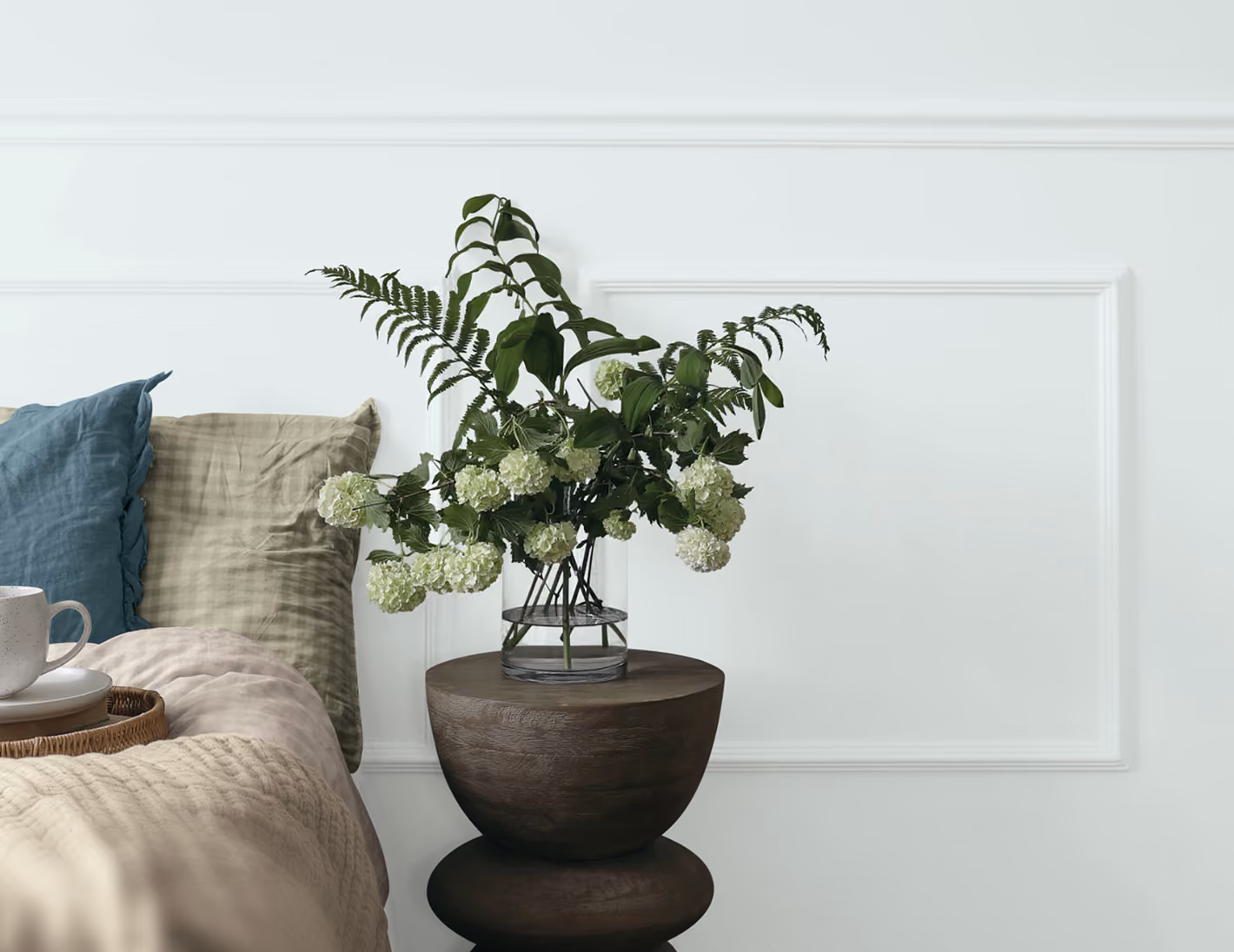 Green flowers in a glass vase on a dark wooden nightstand by a bed