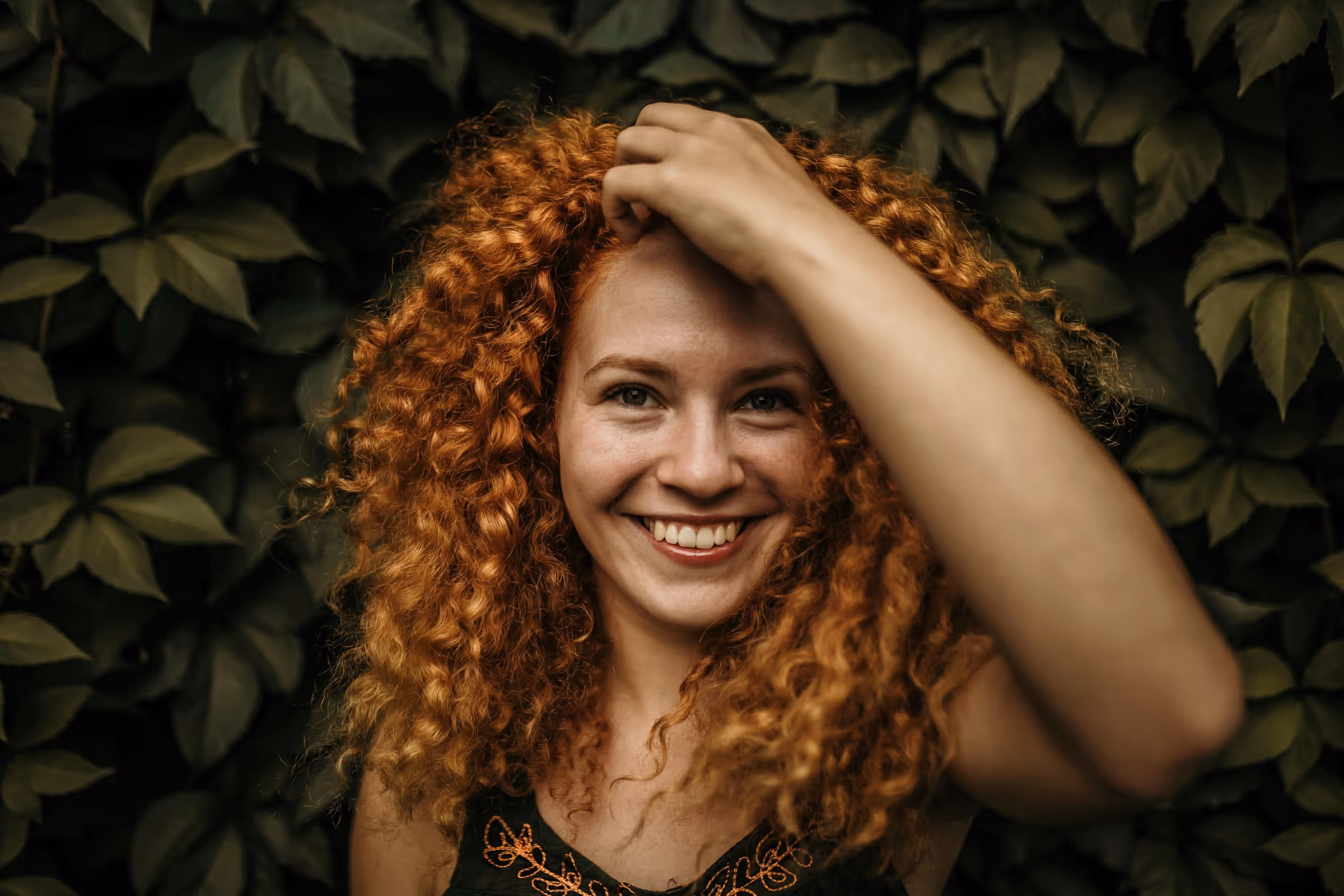 Red-haired woman standing in front of lush vegetation, running her hand through her hair and looking happy, enjoying nature and a joyful moment
