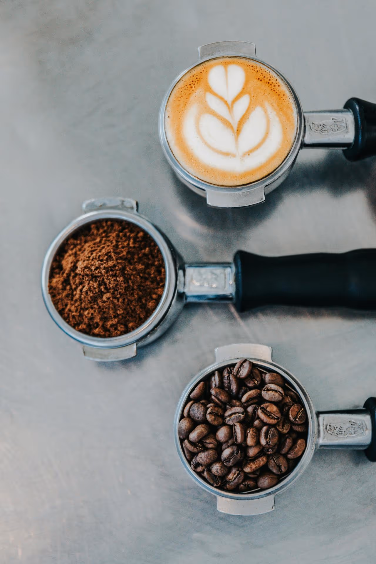 Portafilter trio on a metal surface: top with latte art, middle with ground coffee, bottom with whole beans. Coffee-making process depicted.