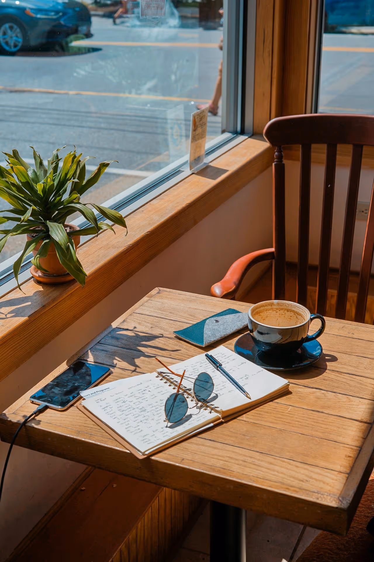 Cozy cafe scene with a wooden table by a window. A notebook, sunglasses, pen, and charging phone are on the table, next to a cup of coffee and a potted plant. Bright, sunny atmosphere.