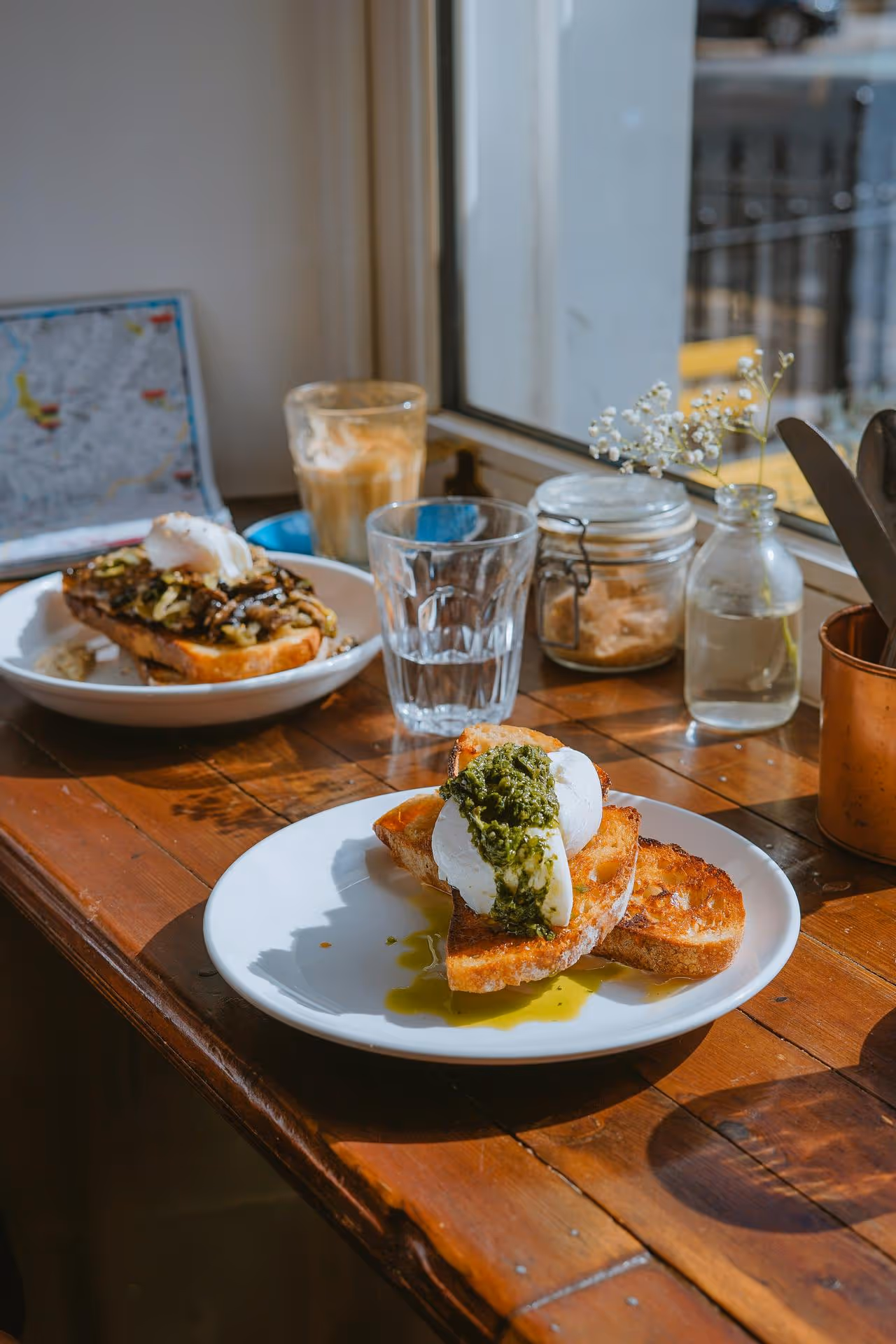 Plates of toast topped with poached eggs and pesto sit on a wooden table by a sunny window. Nearby are a latte, glass, and small vase. Cozy brunch setting.