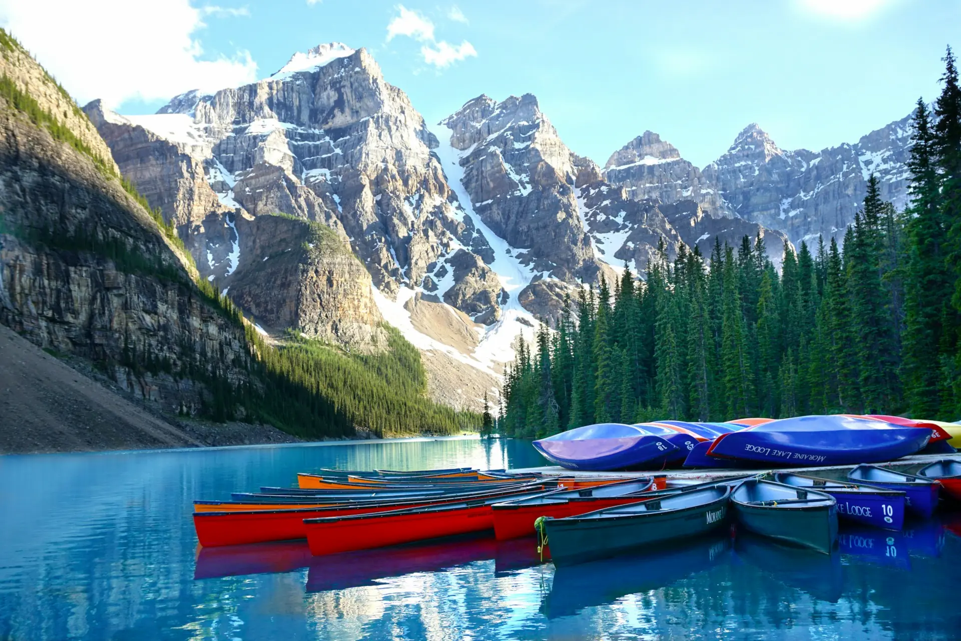 Colorful canoes docked on calm blue Moraine lake with pine trees and snow-capped mountains in the background.