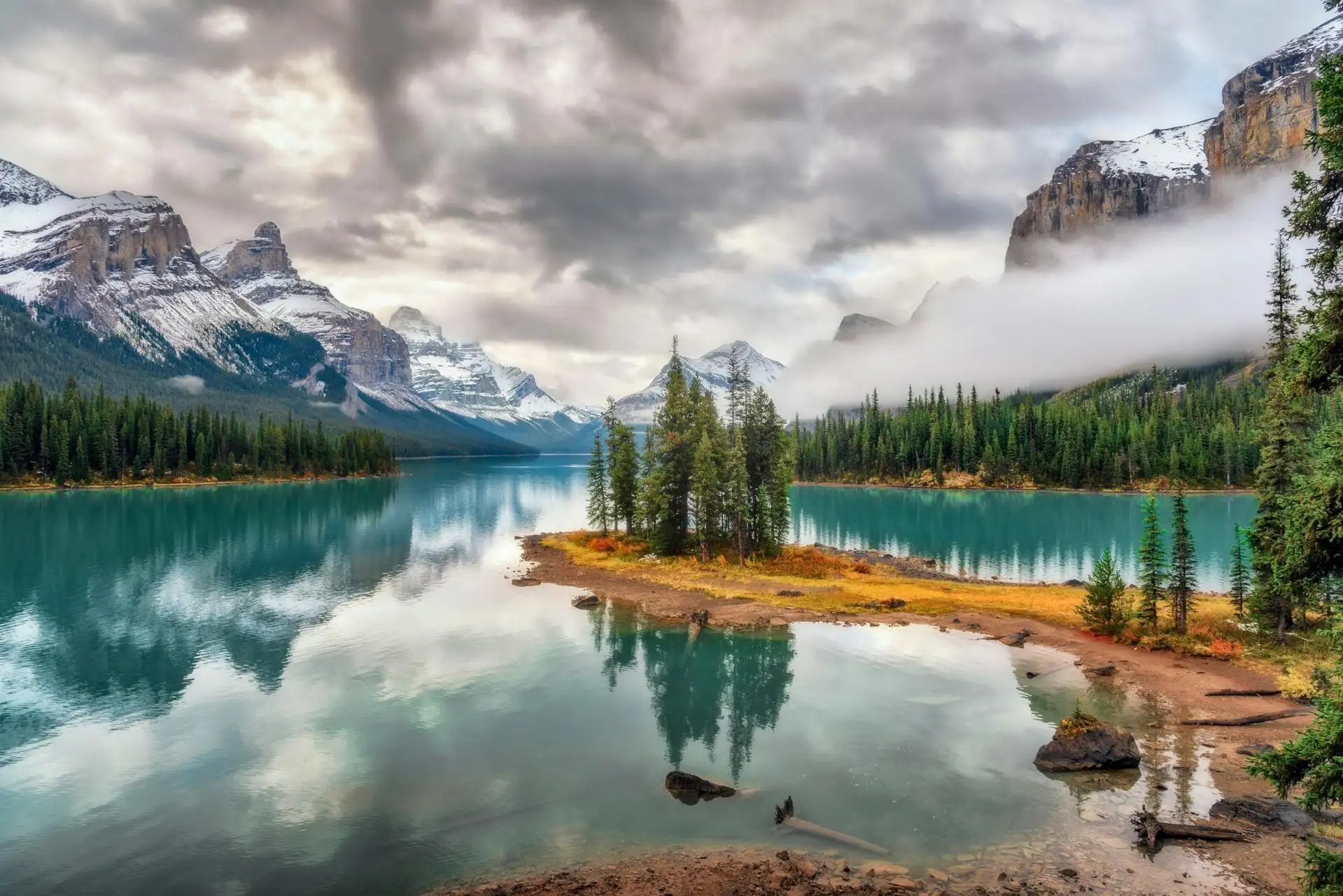 Small forested spirit island in turquoise Maligne Lake surrounded by snow-capped mountains and mist under a cloudy sky.