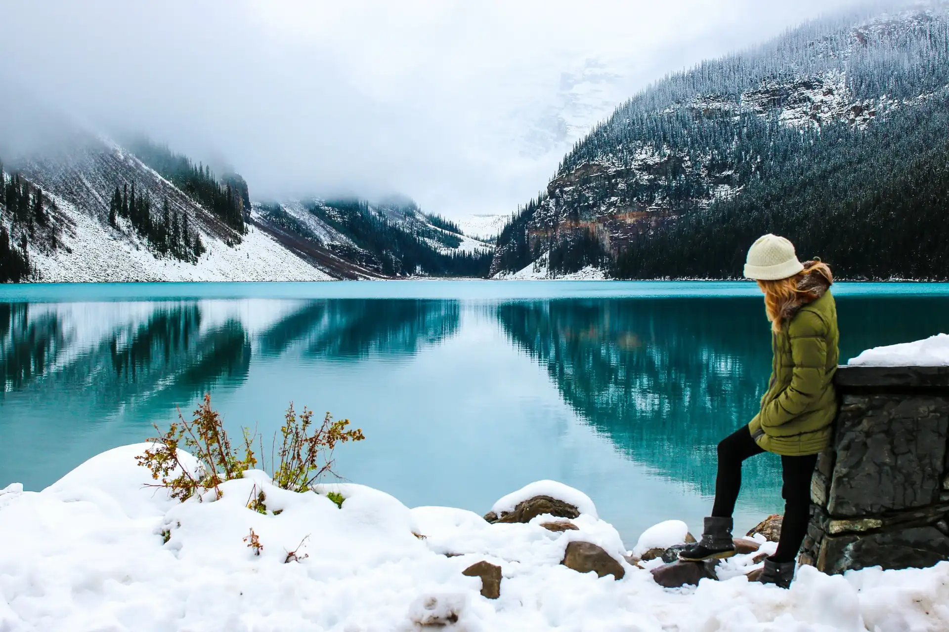 A traveller in green jacket and white hat standing on snowy rocks by the turquoise Lake Louise surrounded by snow-covered mountains and Victoria Mountain hiding in Clouds.