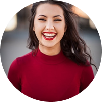 Smiling woman with long dark hair wearing a red top, outdoors with blurred background.