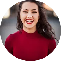 Smiling woman with long dark hair wearing a red top, outdoors with blurred background.