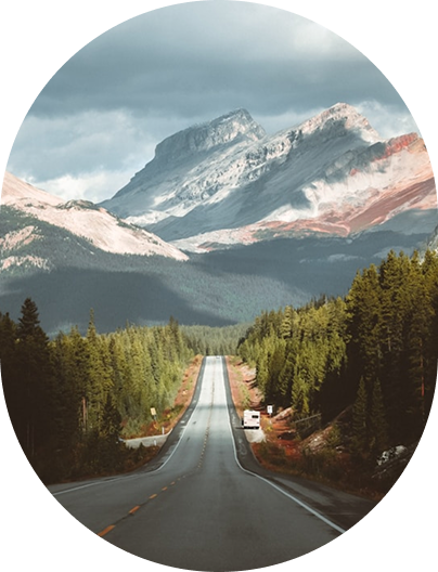 Long straight road leading through dense forest towards large rocky mountains under a cloudy sky.