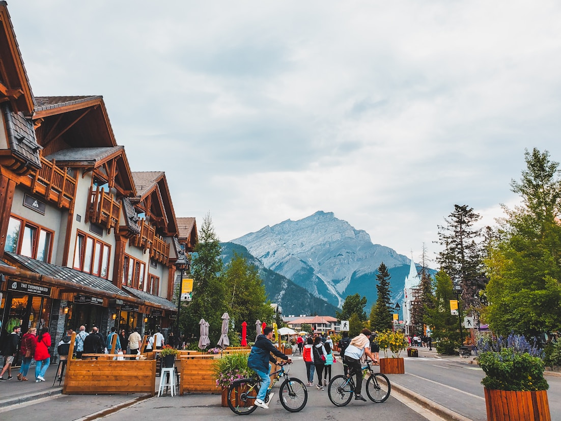 Mountain town street with wooden buildings, people walking and biking, and large mountain in background under cloudy sky.