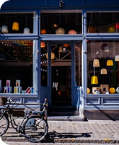 Front of a shop with a glass door and windows displaying colorful hanging lamps, with a bicycle parked outside on the street.