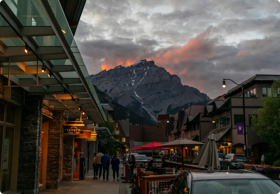 Street scene at dusk with people walking, lit shop signs, parked cars, and a large mountain with a colorful sunset sky in the background.