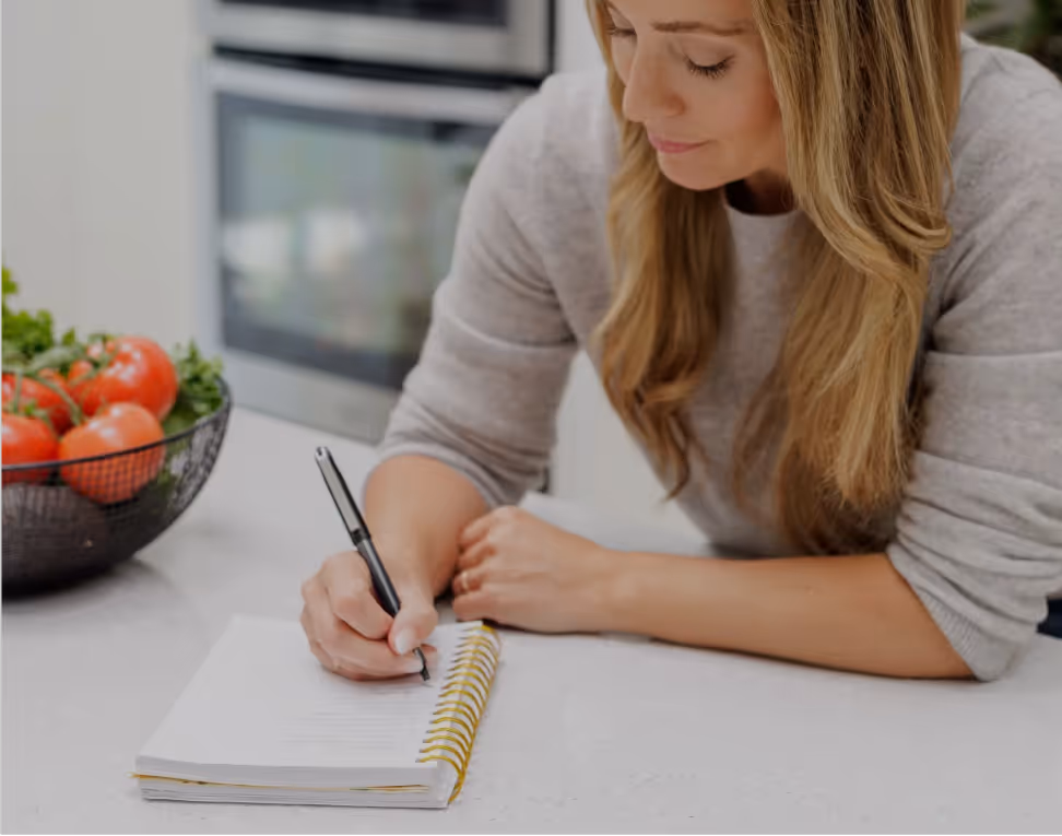 Heather Robertson writing in a spiral notebook at a kitchen counter, with a bowl of tomatoes and vegetables nearby.