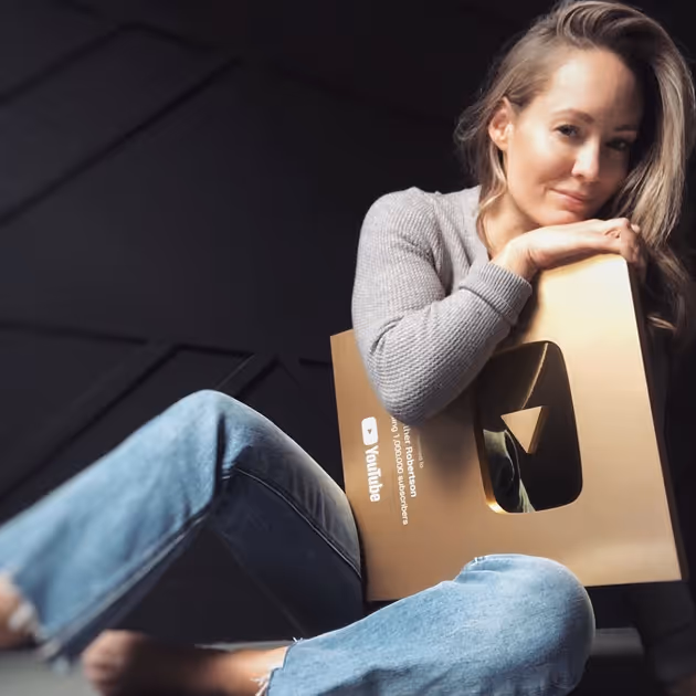 Heather Robertson sitting on the floor holding her YouTube Gold Play Button award, wearing a gray sweater and jeans.