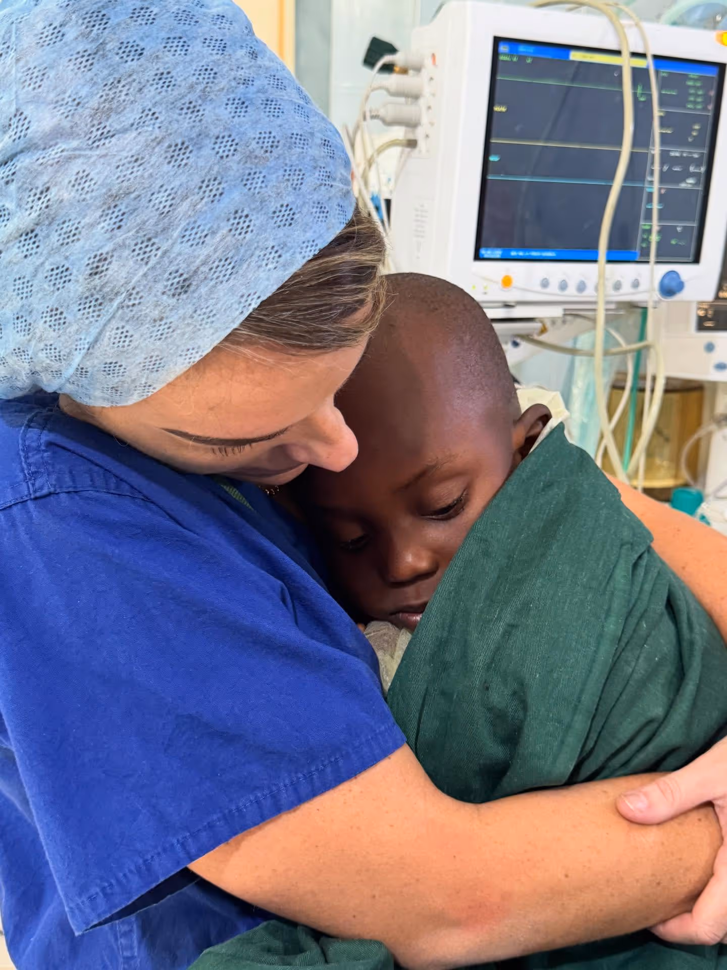 A nurse in blue scrubs gently holds a sleeping child wrapped in a green blanket.