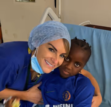A healthcare worker in scrubs smiles and embraces a child sitting on a hospital bed.