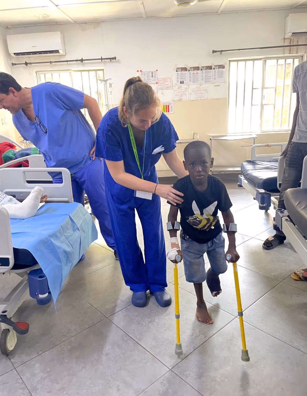 A healthcare worker helps a young boy with crutches walk in a hospital ward.