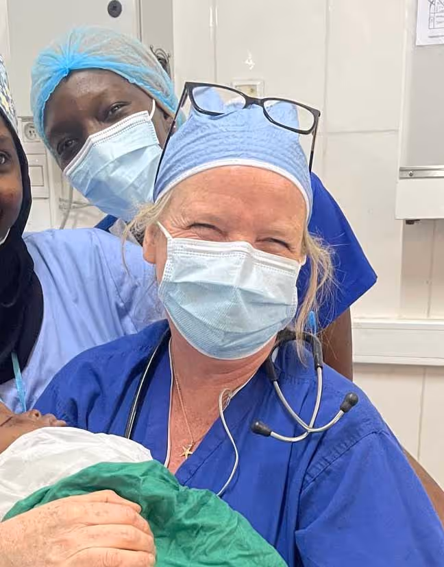 Healthcare workers in blue scrubs and masks smile while holding a wrapped baby.