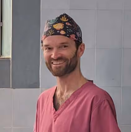 A person in pink scrubs and a colorful cap stands smiling in a tiled room.