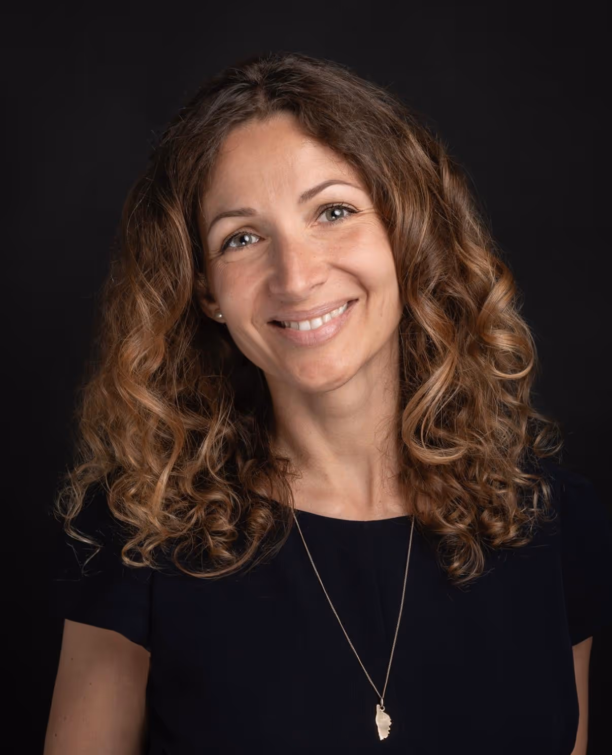 A woman with curly hair smiles at the camera, wearing a dark shirt and necklace.