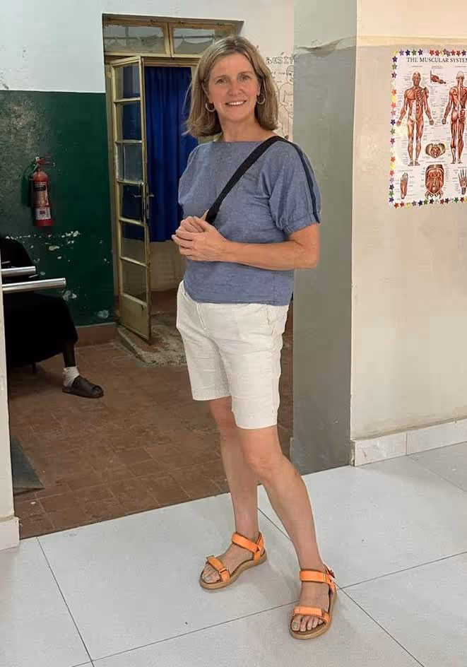 A woman in a blue shirt and white shorts stands smiling in a room with a human anatomy poster.