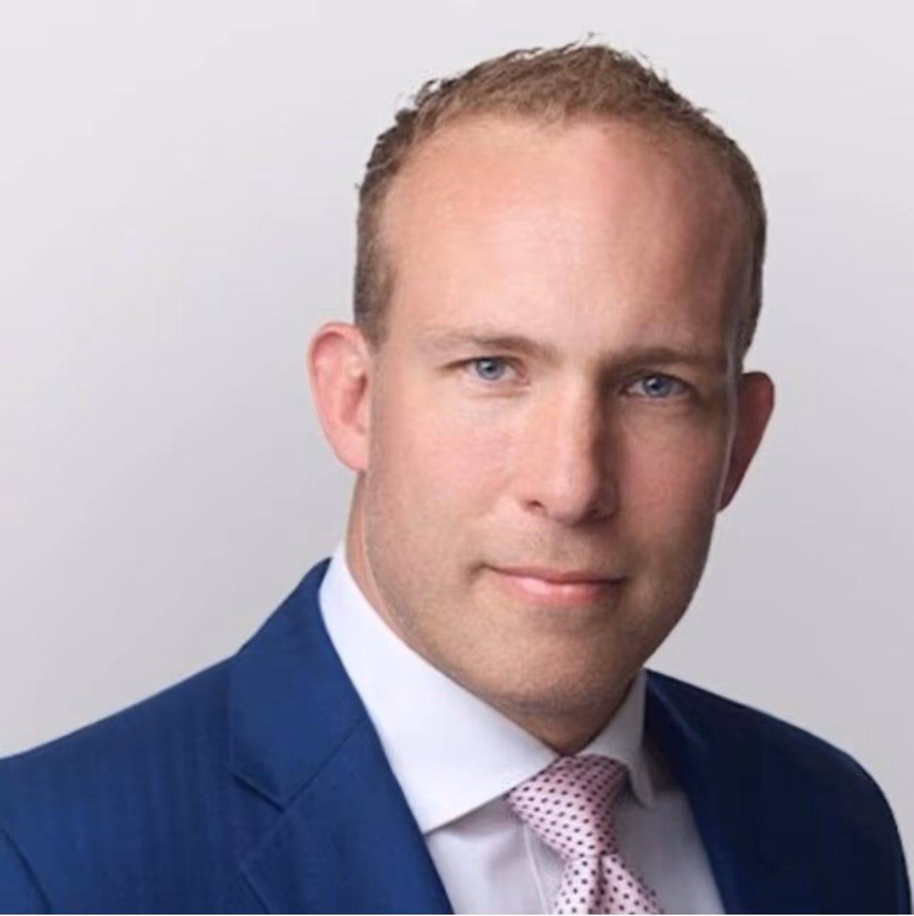 A man wearing a blue suit and pink tie looks directly at the camera.