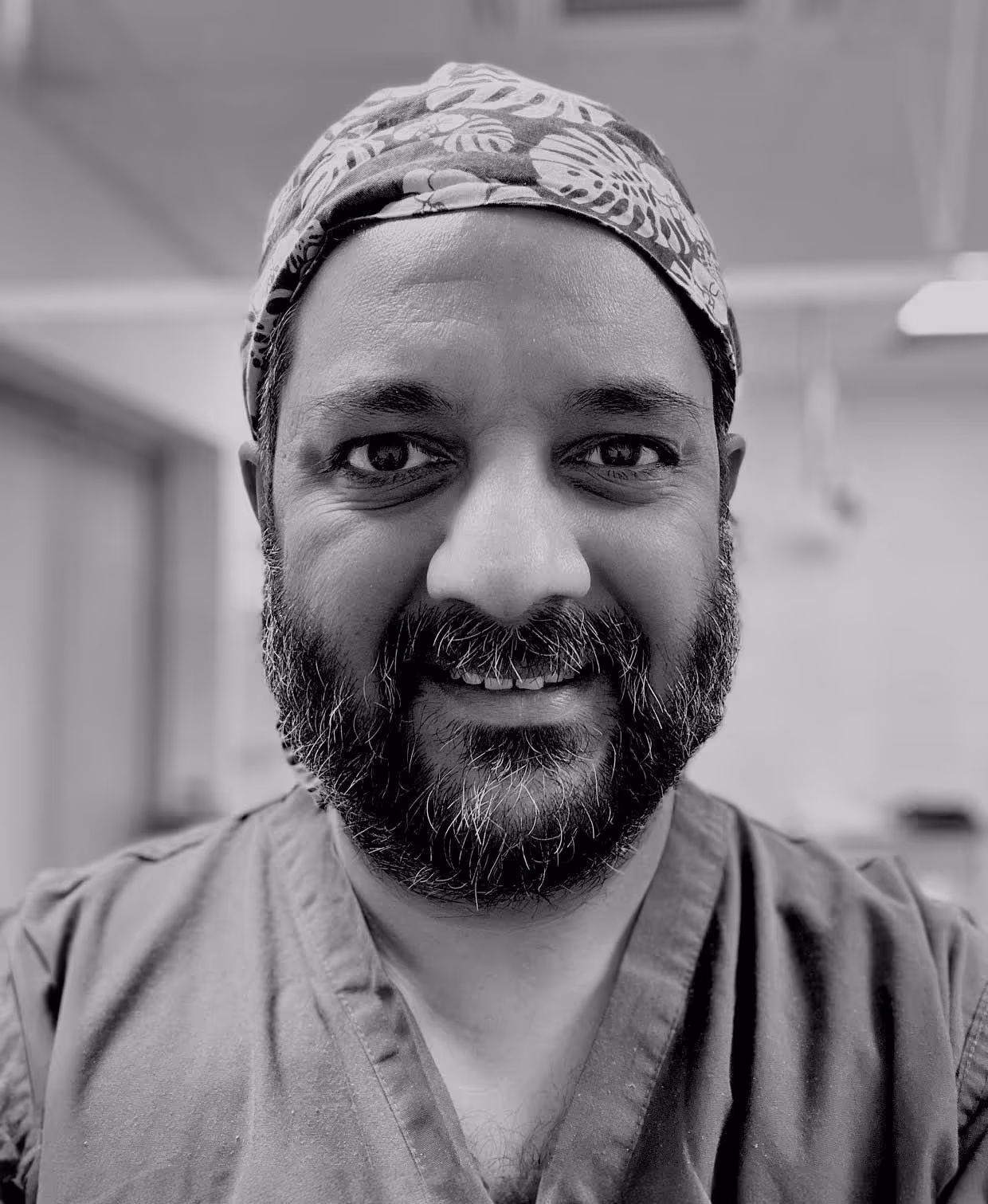 A person wearing scrubs and a patterned headscarf smiles at the camera in a hospital setting.