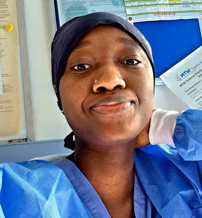 Person wearing a blue surgical gown, headscarf, and nose ring, sitting indoors.