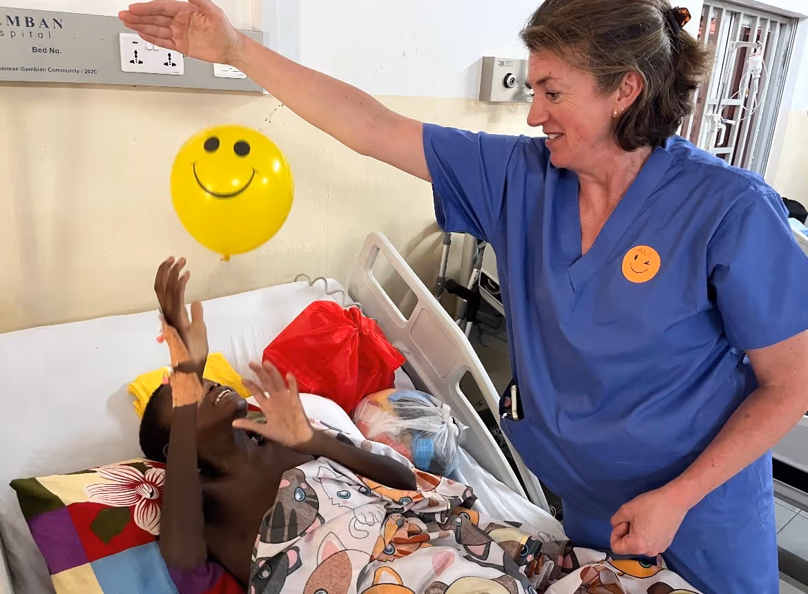 A nurse in blue scrubs plays with a smiling child, using a yellow balloon in a hospital room.