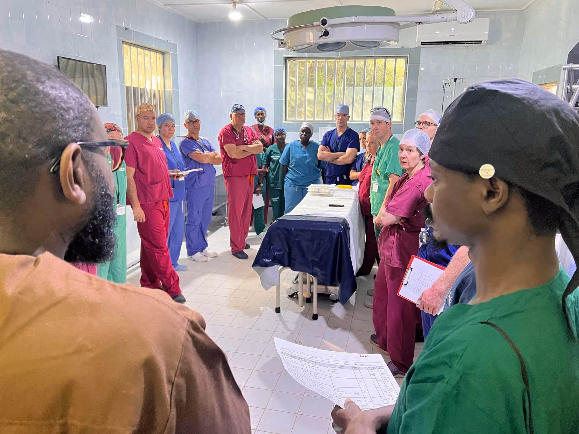 A group of medical staff in scrubs gather around a table in a hospital room.
