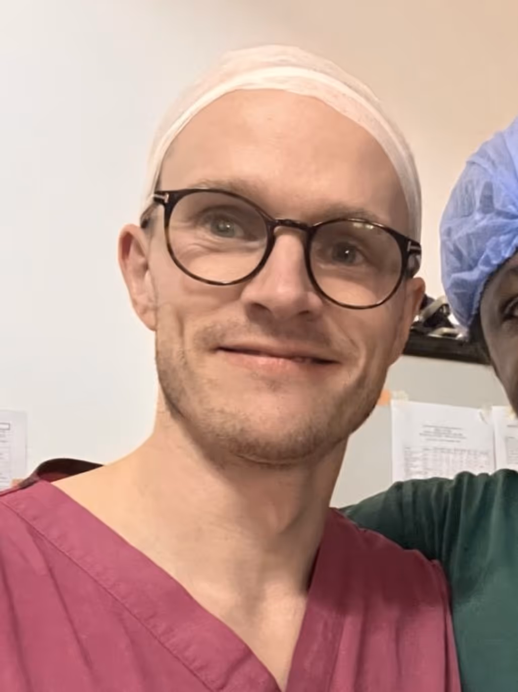 A person wearing glasses and pink scrubs stands smiling, with another person partially visible beside them.