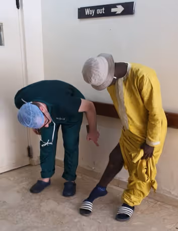 A person in a surgical outfit examines another person's leg in a hallway.
