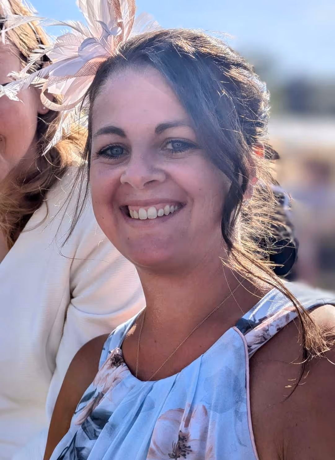 A smiling woman wearing a floral dress and fascinator stands outside in bright sunlight.
