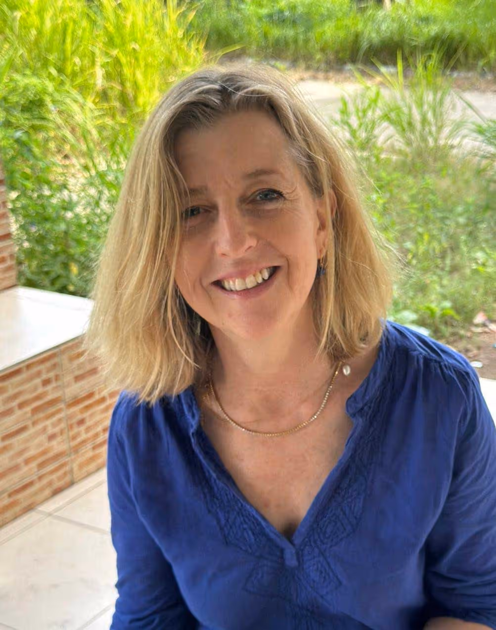 Smiling woman with shoulder-length blonde hair wearing a blue blouse, sitting outside.