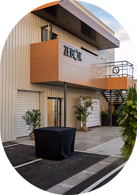 Façade d'un bâtiment moderne avec un balcon en bois marqué 'Zebox' et une table noire devant l'entrée.