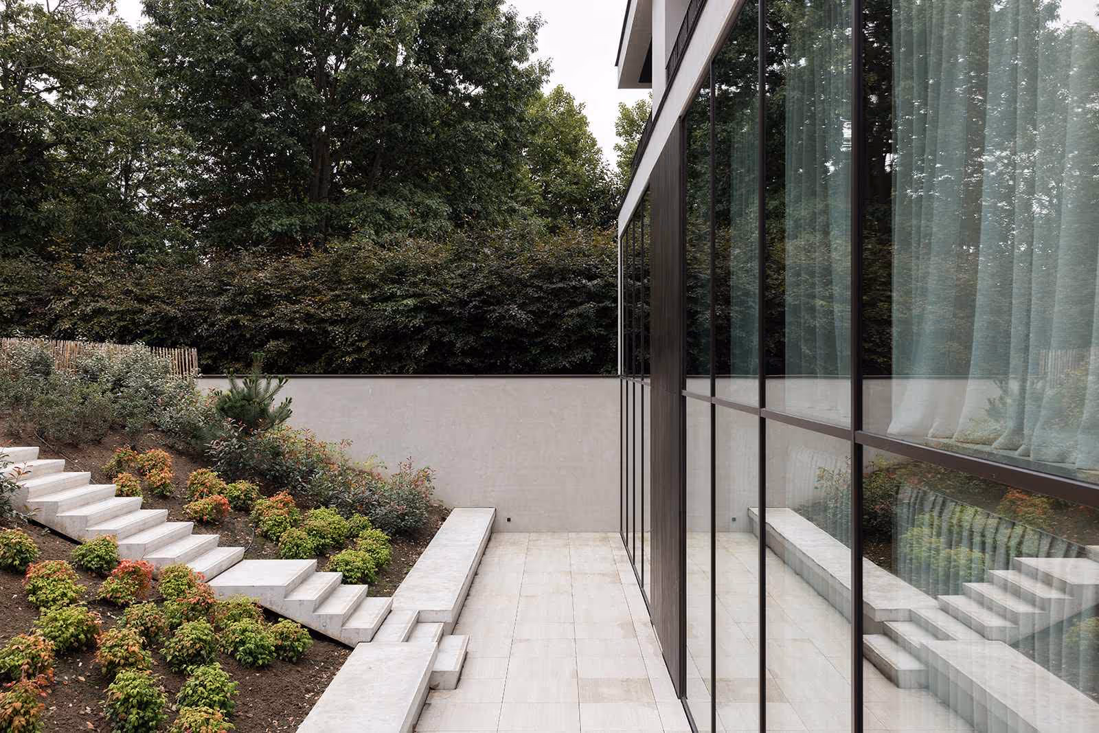 Moderne tuin met witte stenen trappen en een terras naast een glazen gevel met gordijnen, omgeven door groen.